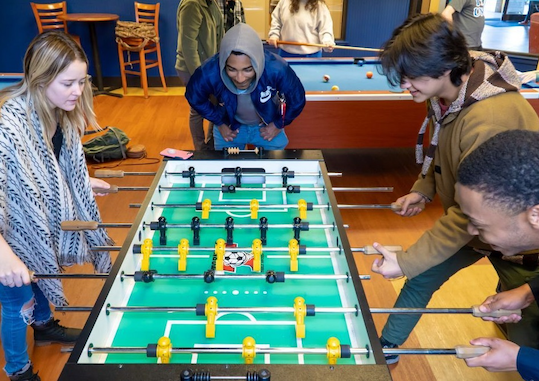 Group of college age students surrounding foos ball table playing a game together of foos ball