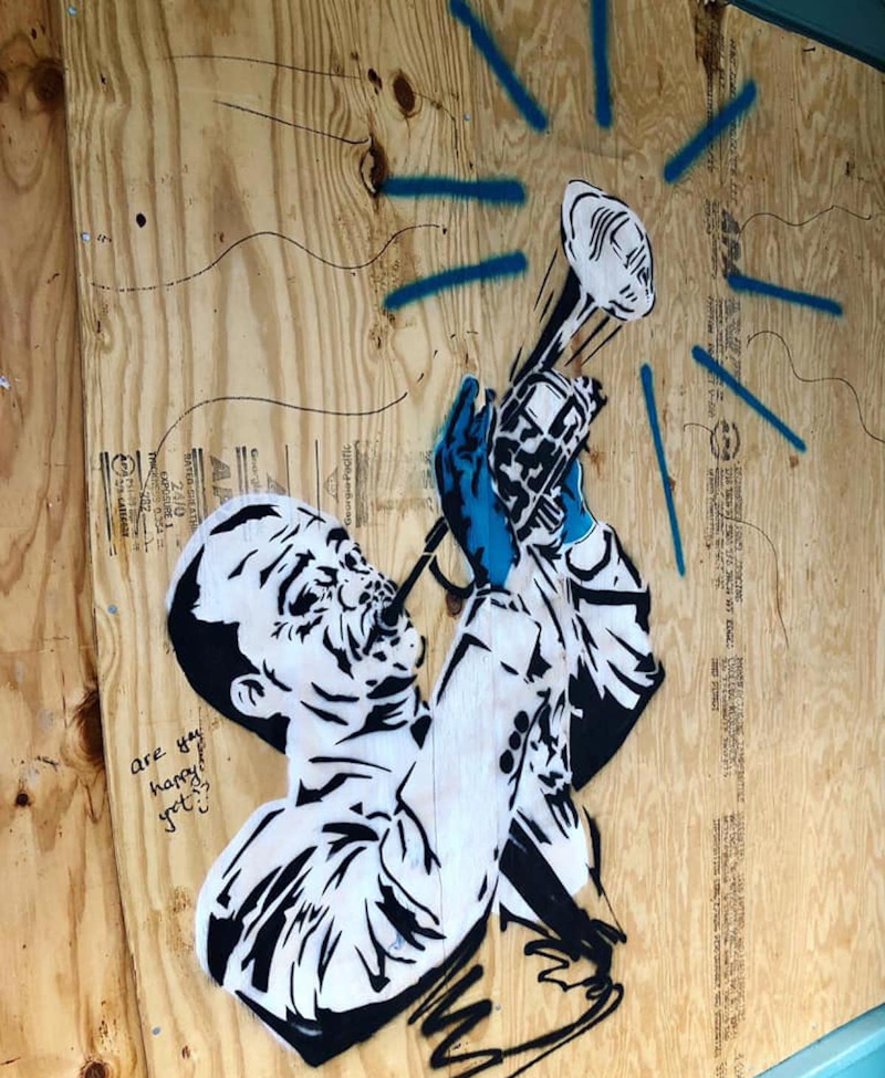 Spray painted image of Louis Armstrong playing trumpet  with a mask over the horn and wearing blue rubber glocves